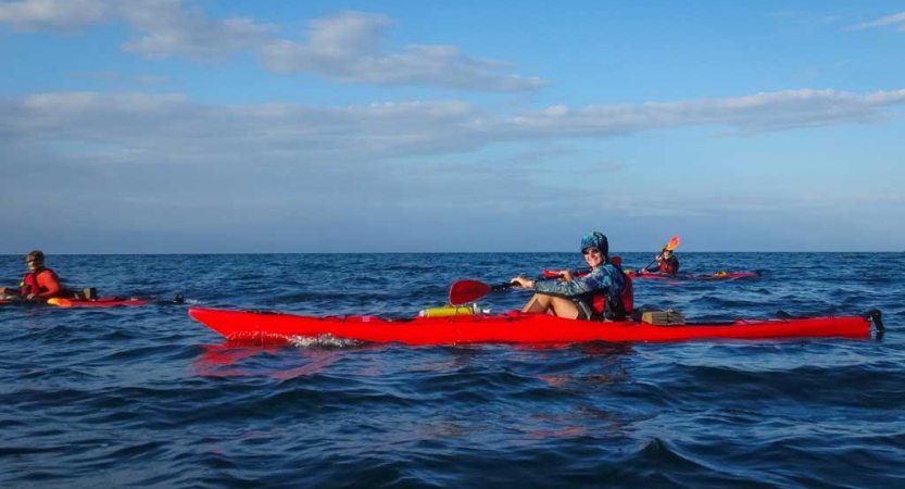 three red kayaks are paddled by outward bound students on a vast blue ocean with a blue sky above.